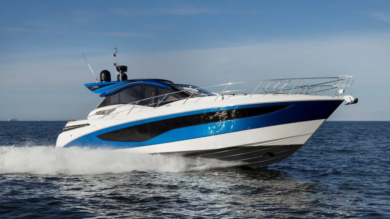 A sleek blue and white motor yacht speeds across the water, creating waves under a clear sky.