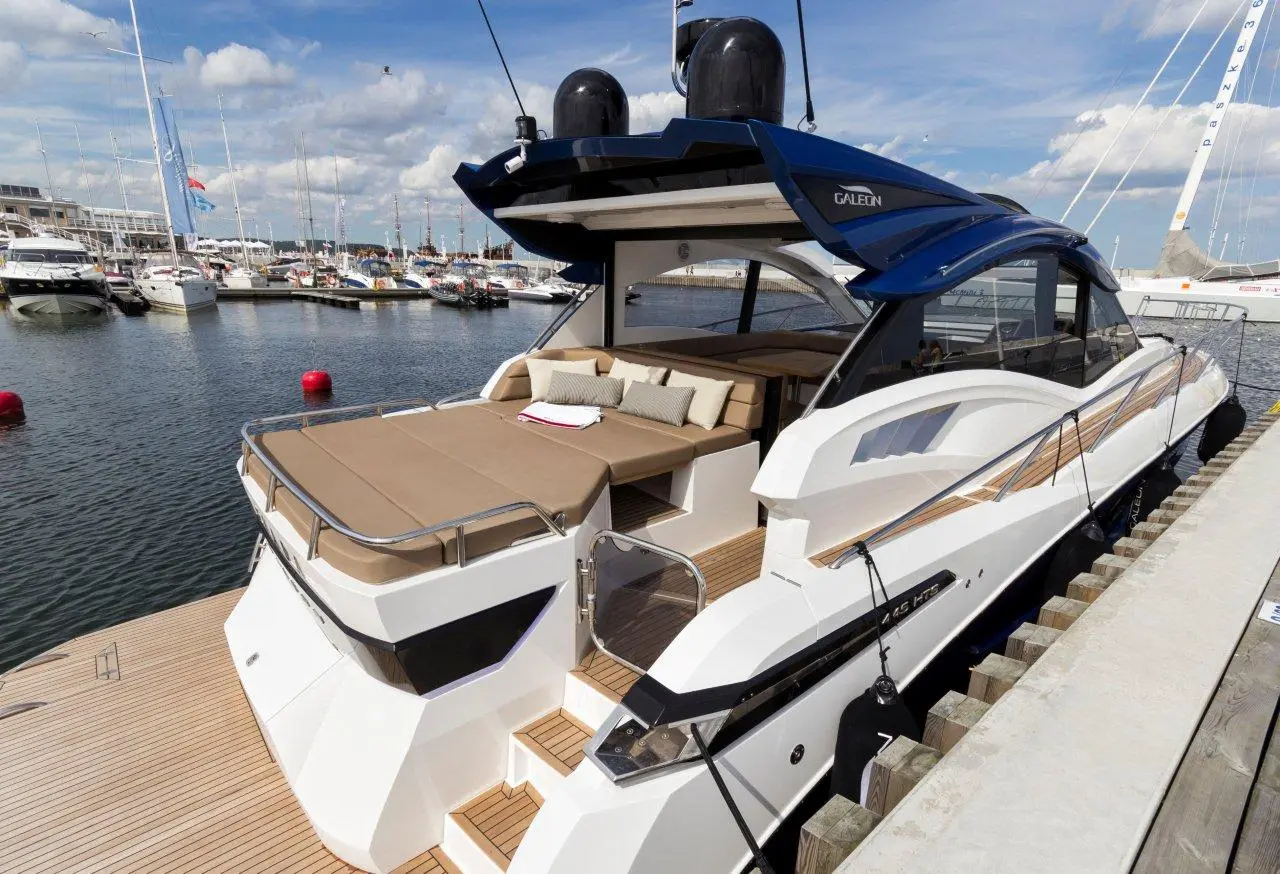 A sleek yacht docked at a marina, featuring a sunbathing area, wooden deck, and surrounding boats on a sunny day.