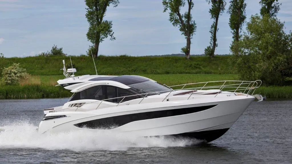 A sleek white motorboat cruising on a river, leaving a wake, with green trees in the background under a clear sky.