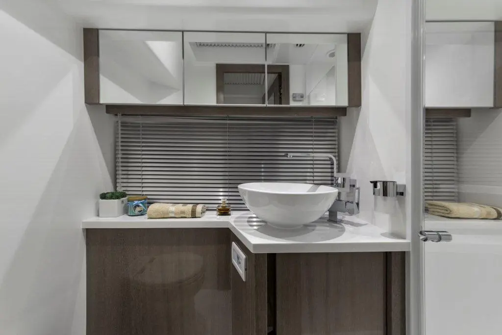 Modern bathroom with a white bowl sink, wooden cabinetry, mirrors, and a window with blinds, featuring towels and plants.
