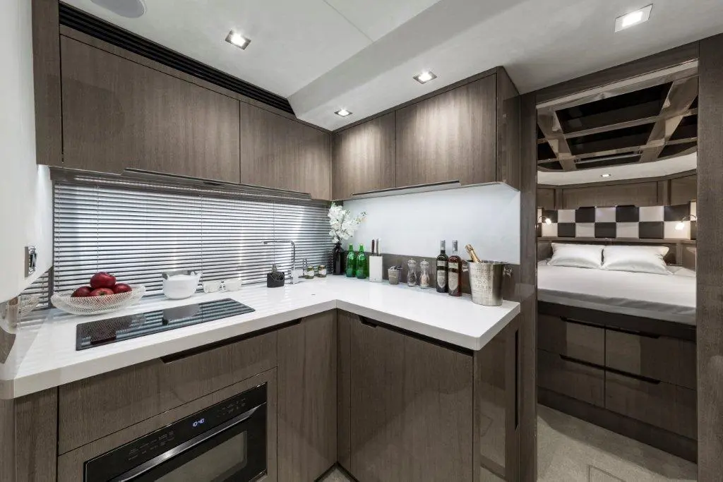Modern kitchen interior with dark wood cabinets, white countertop, stove, wine bottles, and a bedroom visible in the background.
