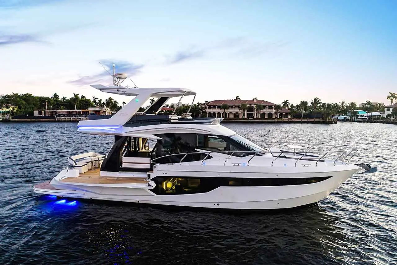 Luxury motor yacht docked on water, featuring sleek design and illuminated underbelly, with upscale waterfront homes in the background.