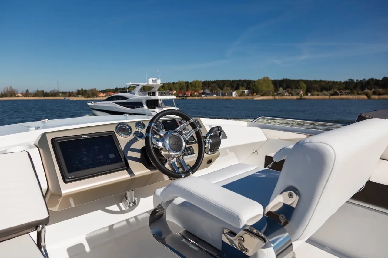 Boat cockpit with steering wheel, display panel, and white seat overlooking water with another boat in the background.