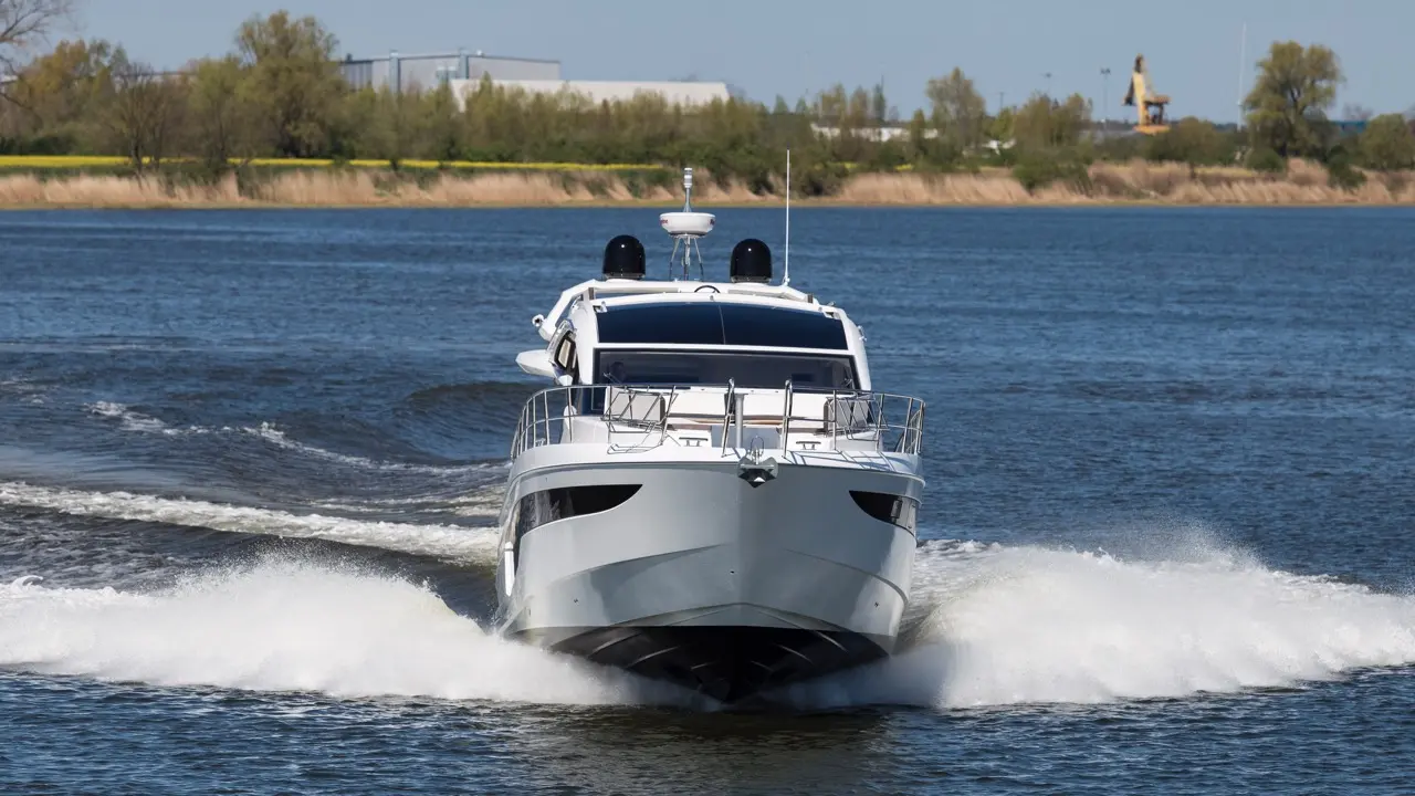 A sleek motor yacht navigates through water, creating a wake, with trees and industrial buildings in the background.