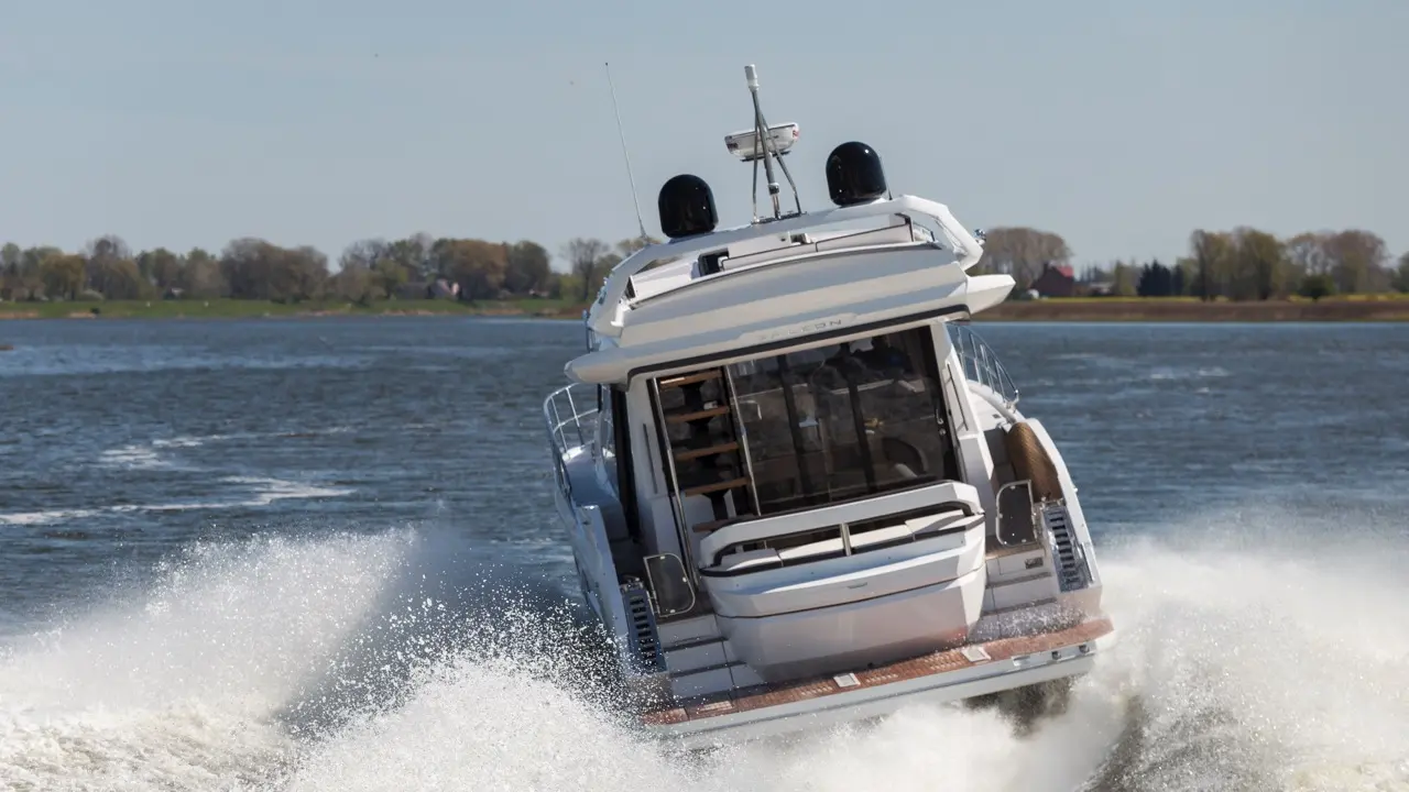 A luxury motorboat lifts out of the water while creating a spray of waves, in a scenic river setting on a sunny day.