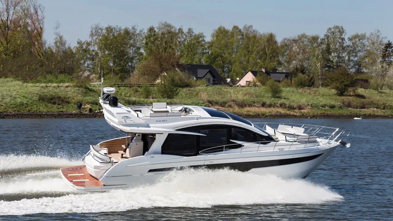 Luxury motor yacht sailing on a calm river, creating white waves, surrounded by green trees and residential homes in the background.