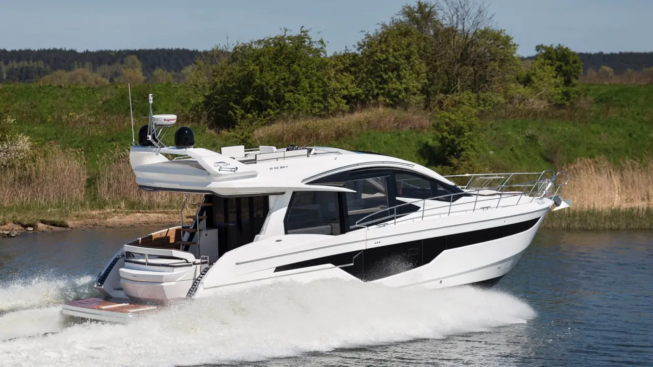 A sleek white motor yacht glides through water, creating a spray, surrounded by lush greenery under a clear blue sky.