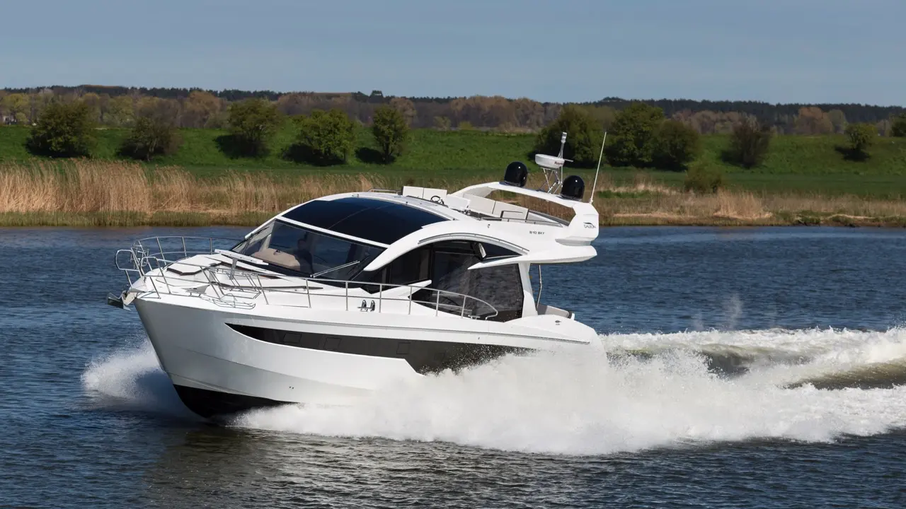 A sleek white motor yacht speeds across a river, creating splashes, with green landscapes and trees in the background.