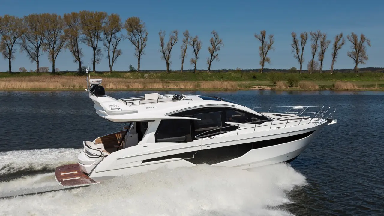 A sleek motor yacht cruises on calm water, leaving a white wake, with a row of trees in the background under a clear blue sky.