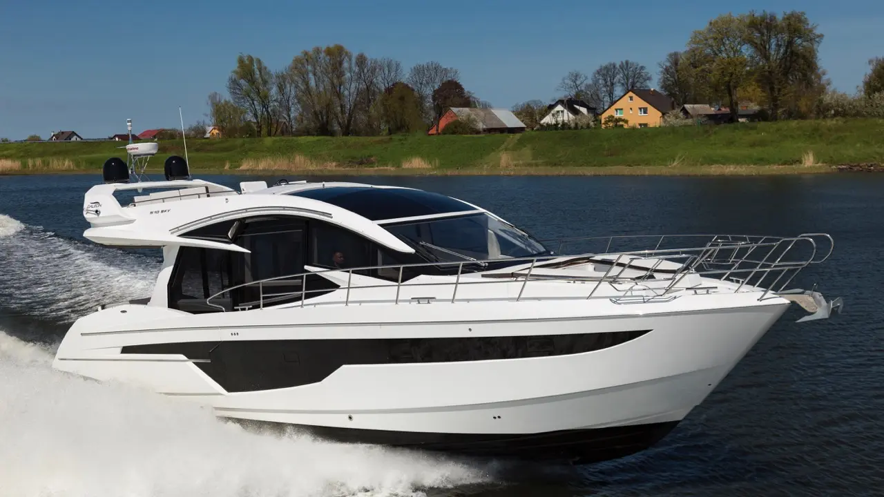A sleek white motor yacht cruises through calm waters, leaving a wake, with greenery and houses visible in the background.