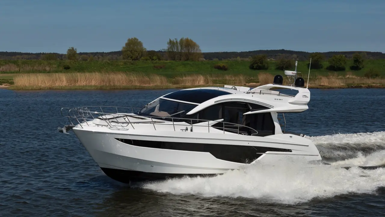 A sleek white motorboat glides through calm waters, with green landscape and trees in the background under a clear blue sky.