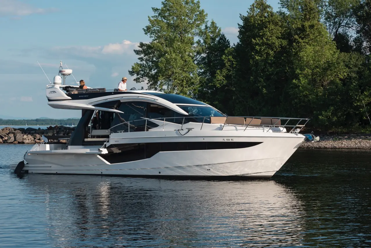 A sleek white yacht with outdoor seating, gliding on calm waters, surrounded by trees under a clear blue sky.