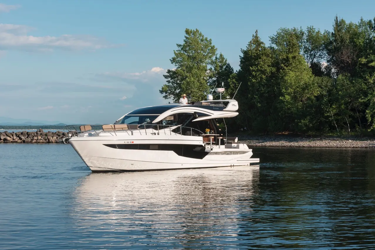A sleek white motor yacht with a captain at the helm, gliding across calm water near a rocky shore and lush trees.