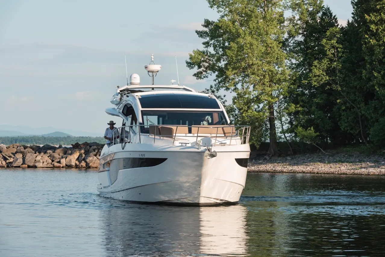 A luxury motor yacht glides through calm waters near a rocky shore, surrounded by green trees under a clear sky.
