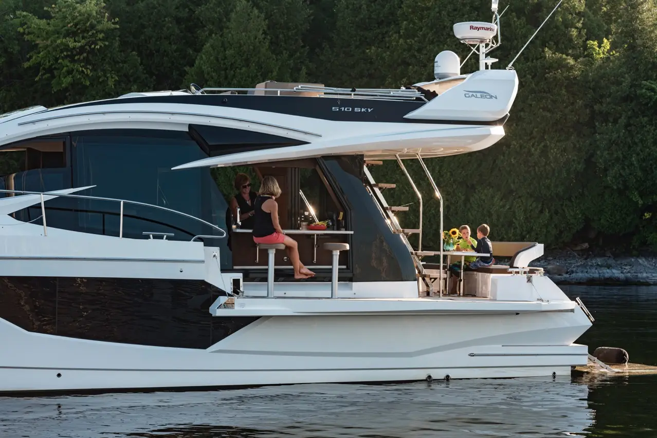 A luxurious Galeon S510 Sky yacht, featuring outdoor seating, two women, a child, and sunflowers surrounded by lush greenery.