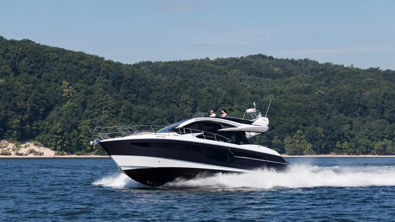 A sleek black and white yacht glides across blue water, with two people on deck, surrounded by green hills.