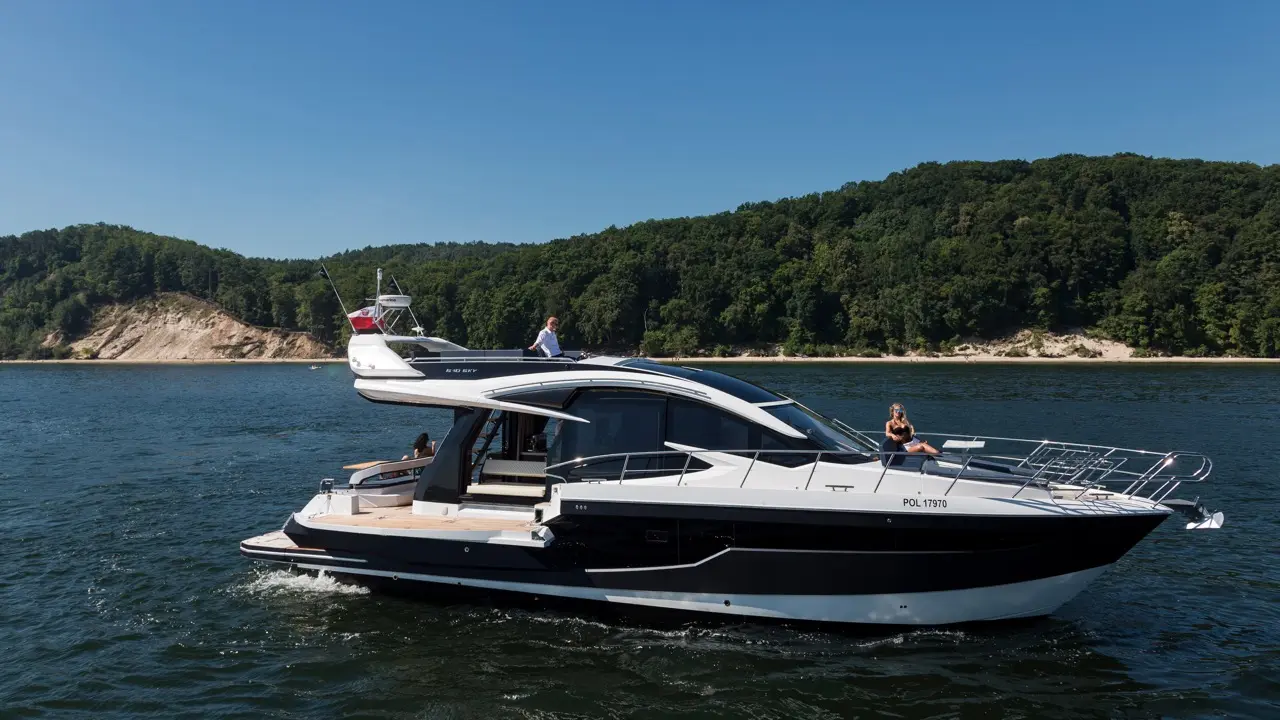 A sleek black and white yacht on a lake, with two people onboard; one is standing and the other sitting, enjoying the sun.