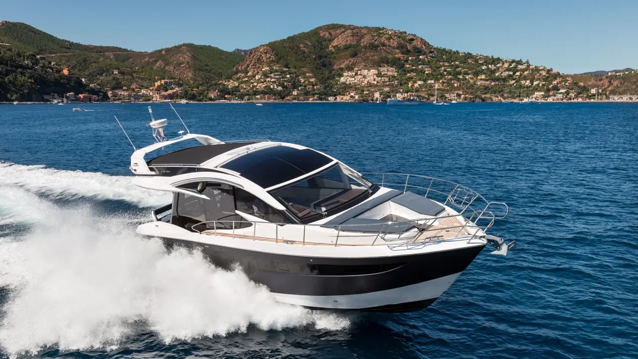 A sleek motorboat speeds through blue waters, leaving a white wake, with green hills and coastal homes in the background.