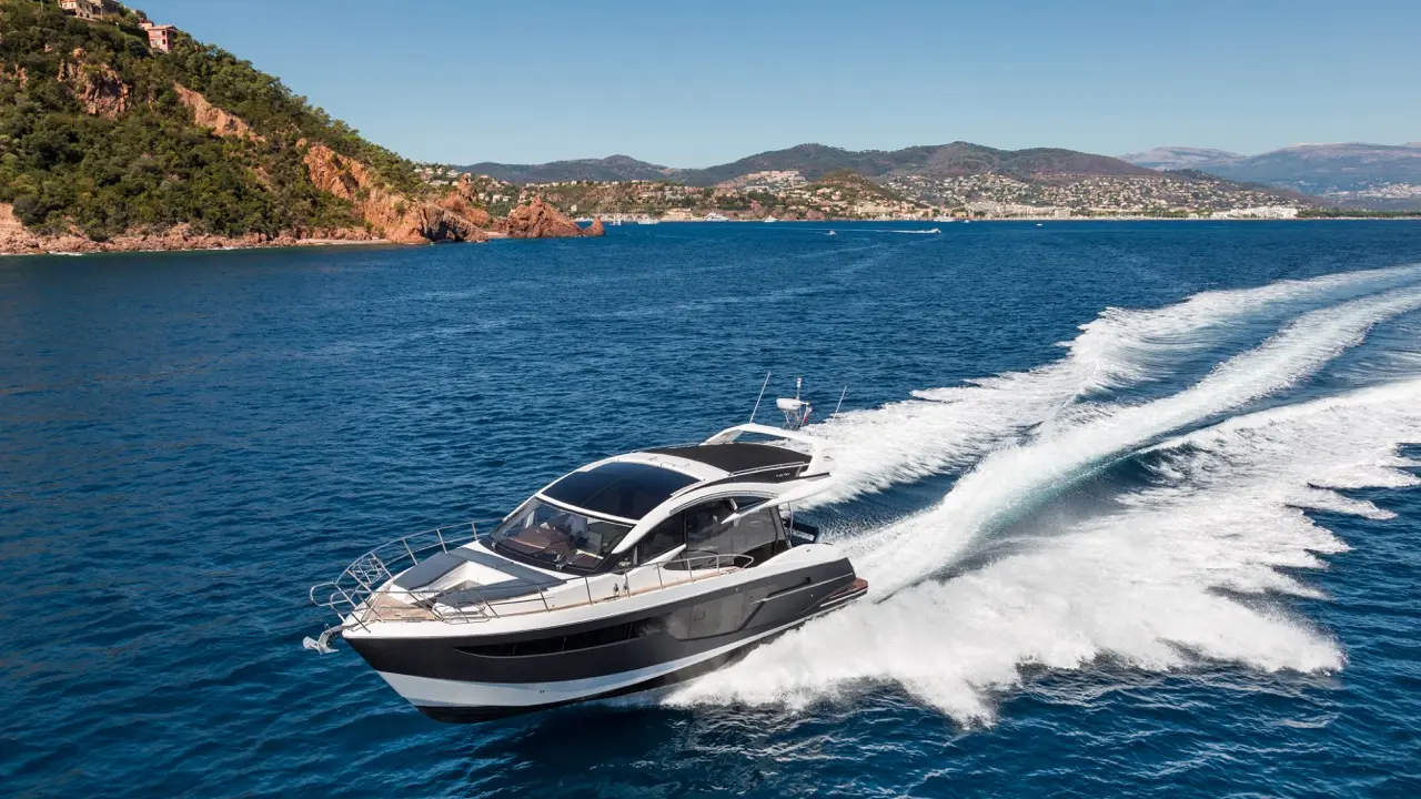 A sleek motorboat speeds through turquoise waters, leaving a white wake, with green hills and a coastal town in the background.
