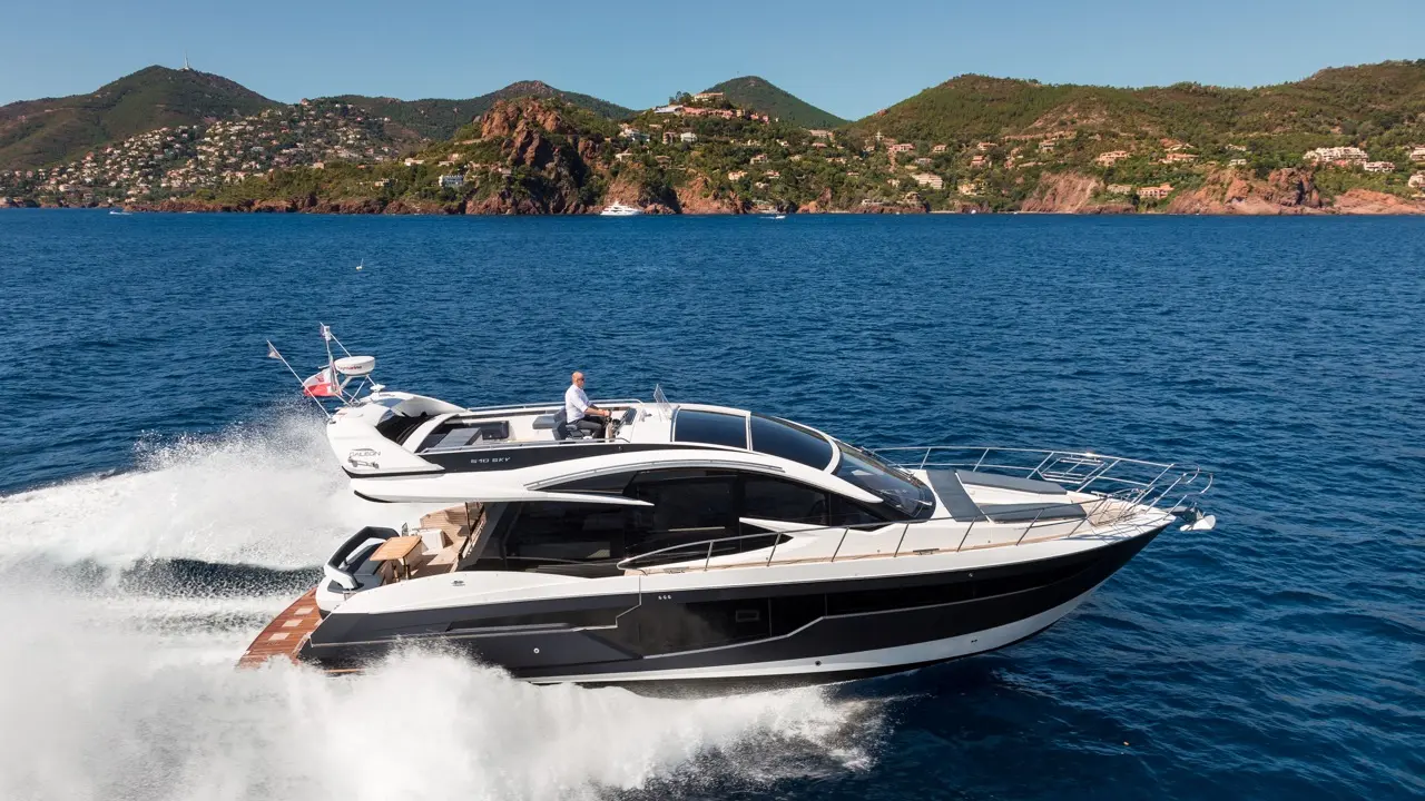 A luxury yacht speeds through deep blue water, with a man at the helm and scenic hills in the background.