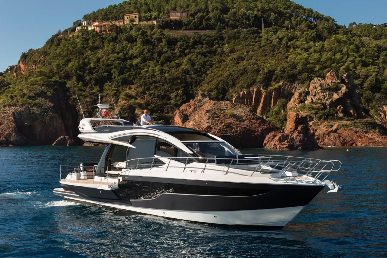 A sleek motor yacht cruises calmly on clear blue water, with rocky cliffs and lush greenery in the background.