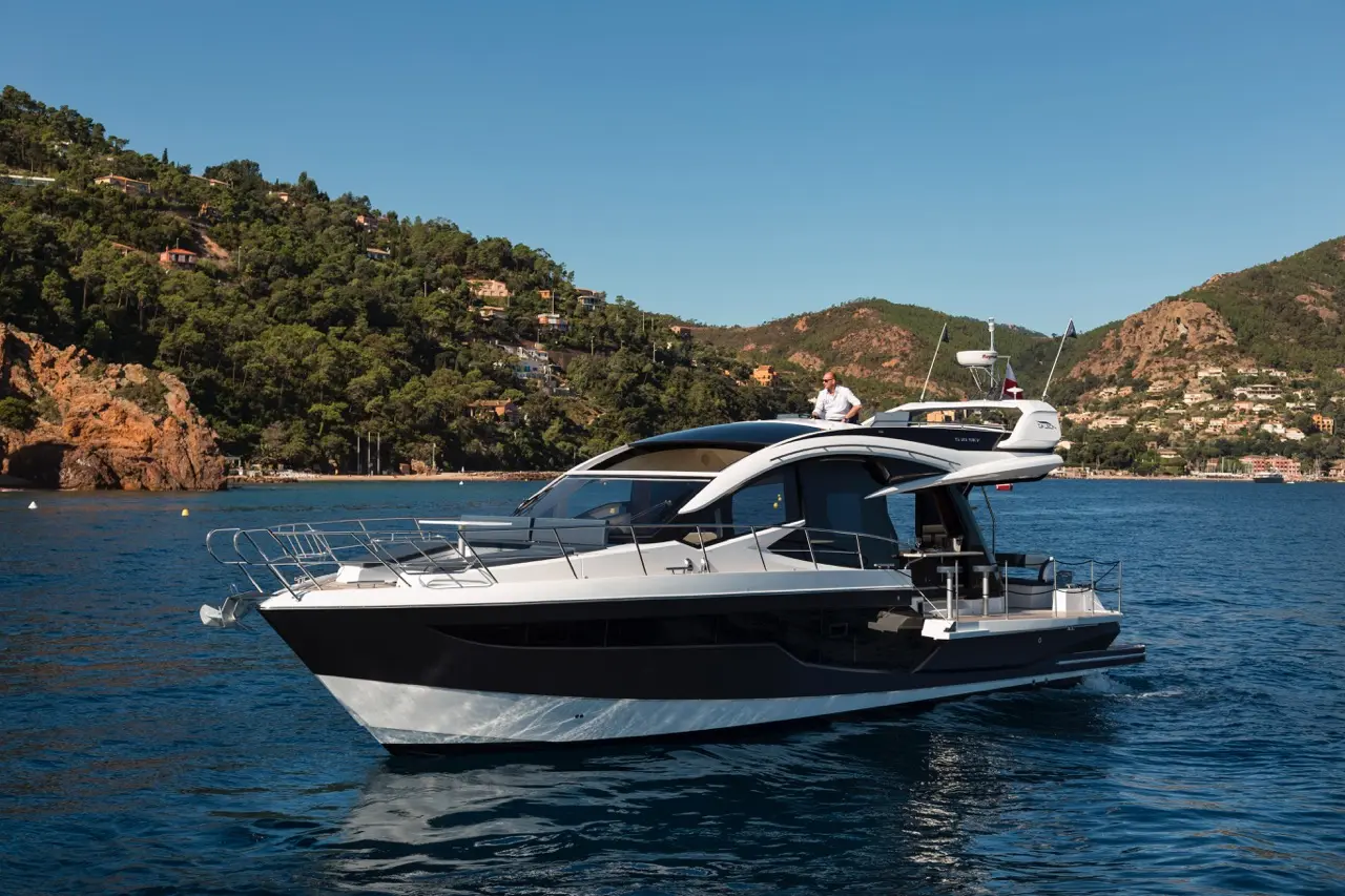 A sleek modern yacht with a man on the upper deck, anchored near a lush hillside and coastal homes under a clear blue sky.