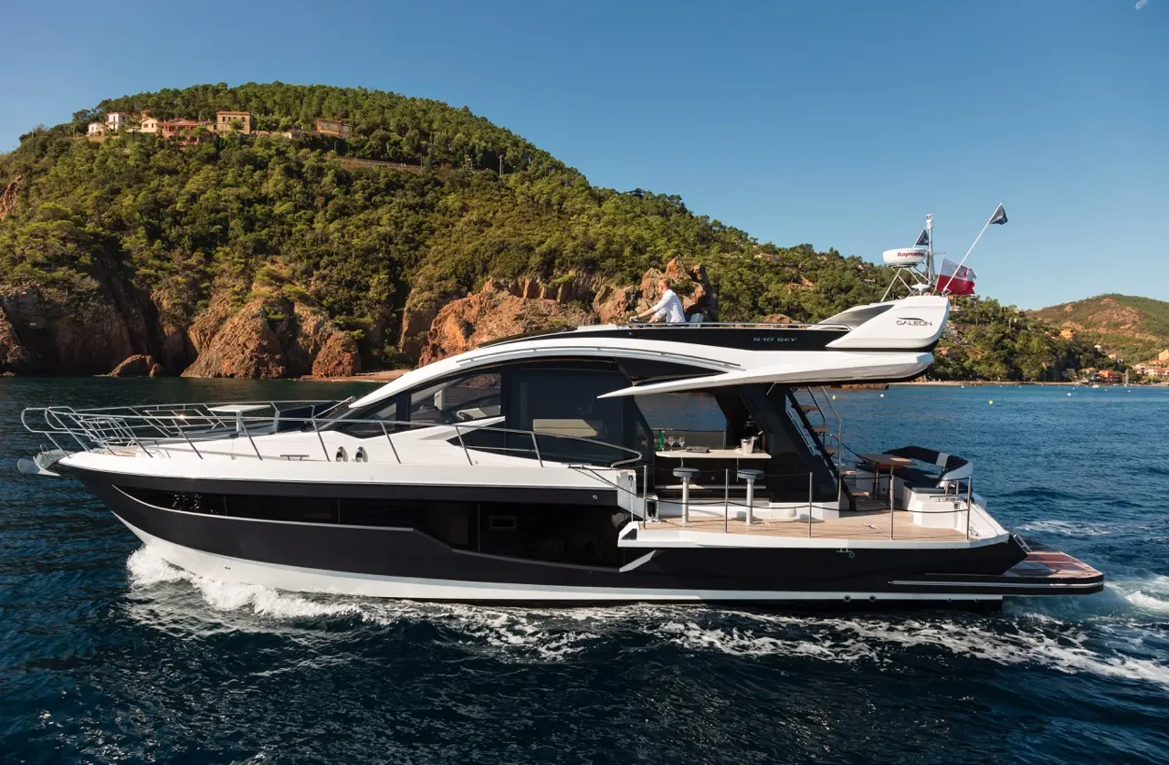 Luxury yacht cruising on calm waters, with a person at the helm and lush hills in the background.