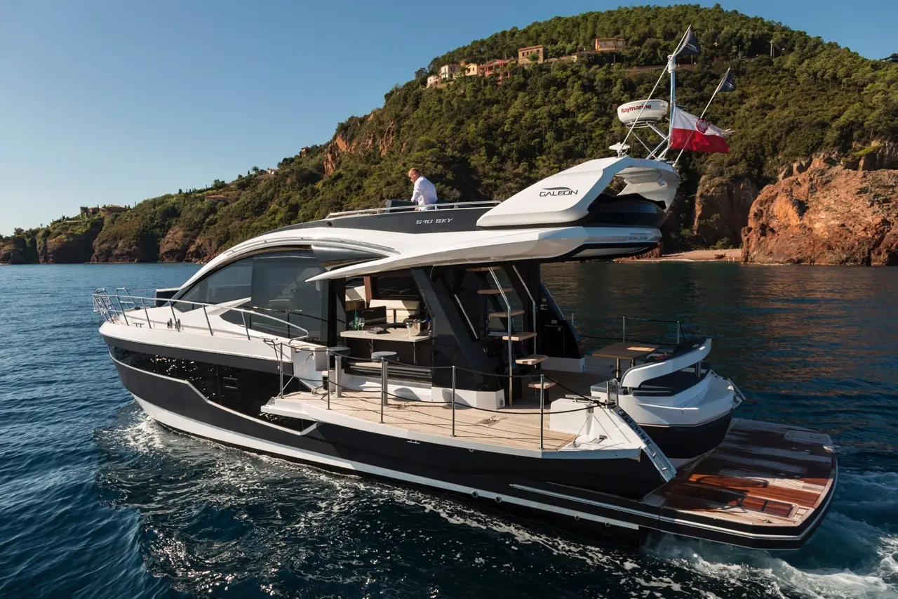 Galeon 510 Sky yacht cruising on blue waters with a man on deck, surrounded by lush green hills and rocky cliffs.