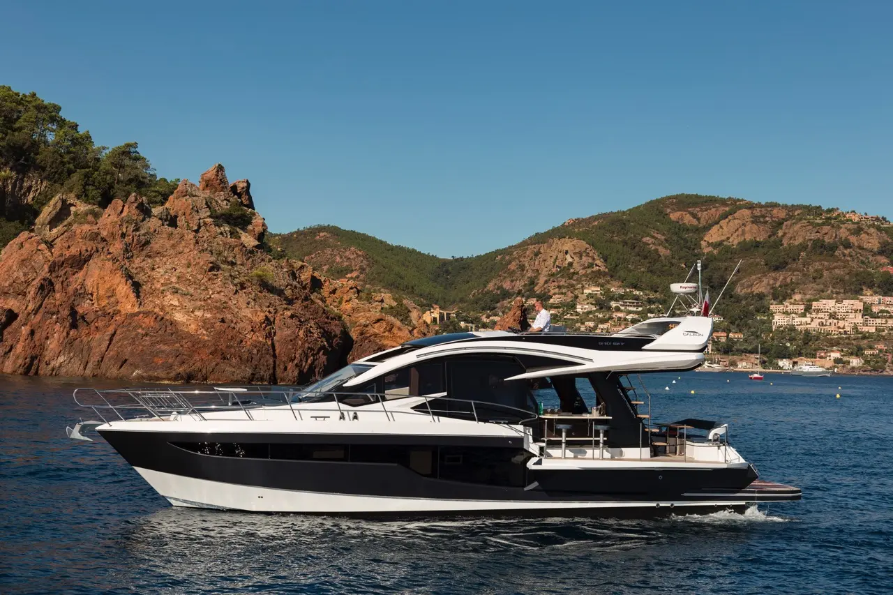 A sleek motor yacht gliding on blue water near rocky cliffs and green hills, with a person relaxing on the top deck.