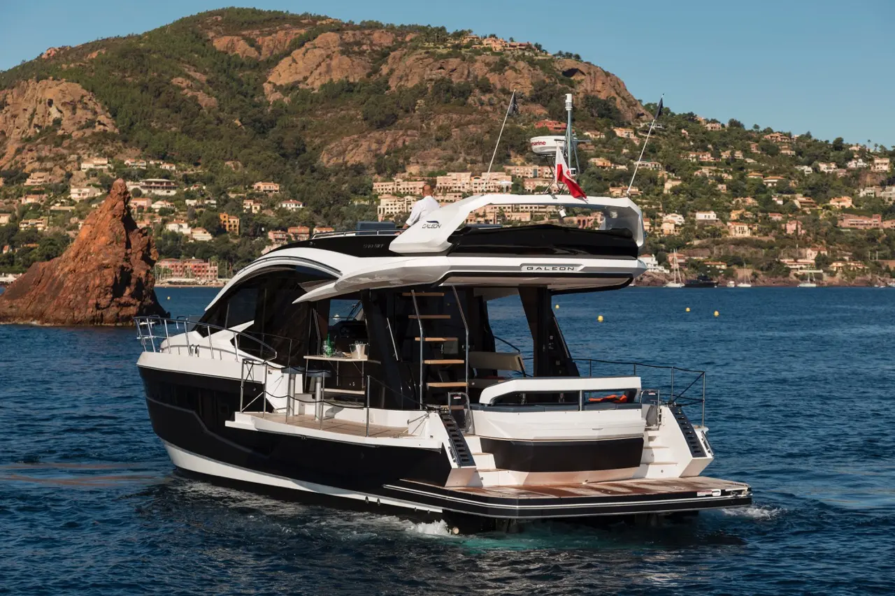A luxury yacht named Galeon on water, with a coastal backdrop of colorful homes and steep hills, under clear blue skies.