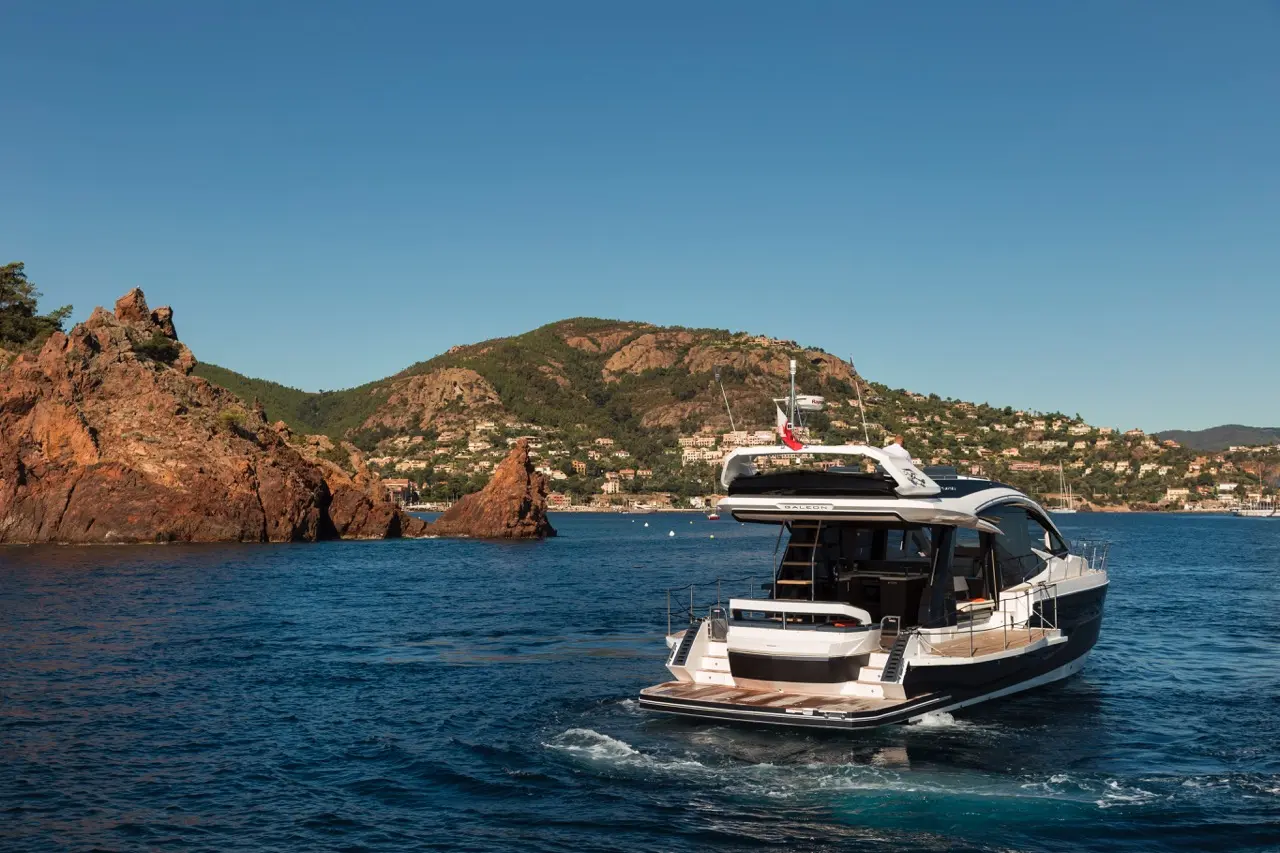 Luxury yacht navigating blue waters near rocky formations and a scenic coastal village under a clear sky.