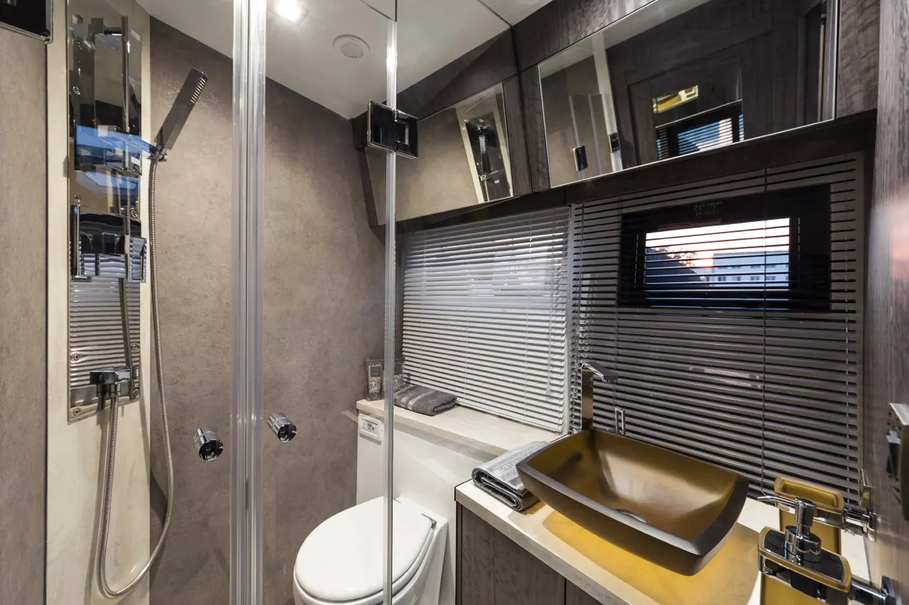 Modern bathroom with a glass shower, toilet, stylish sink, mirror, and window with blinds, featuring sleek gray and gold design.
