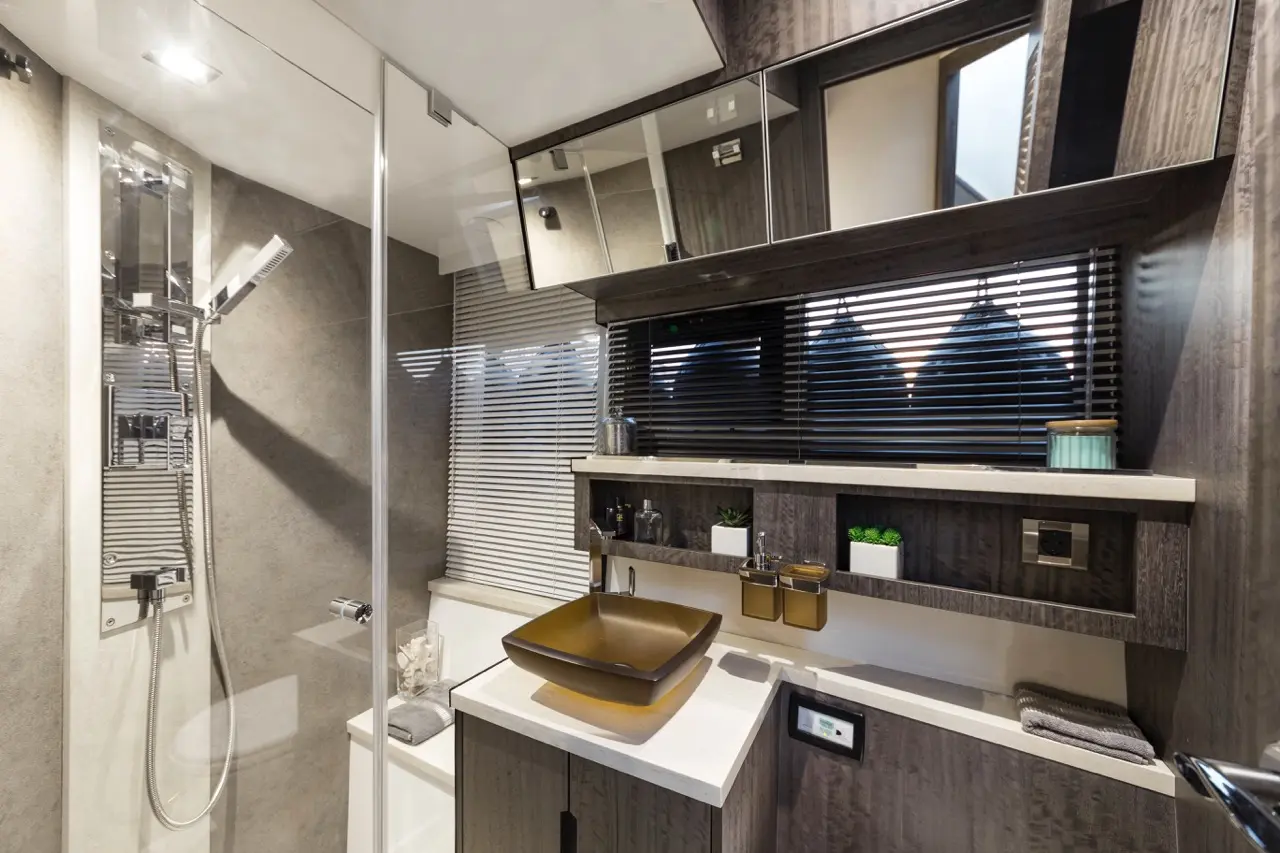 Modern bathroom featuring a glass shower, brown basin, and wooden shelves with plants and toiletries against a gray backdrop.