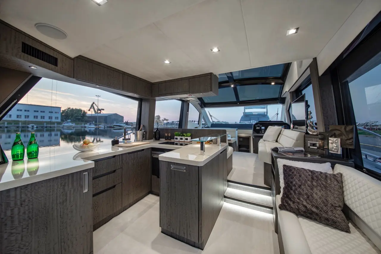 Modern yacht interior featuring a sleek kitchen area, plush seating, and large windows with views of the harbor at sunset.