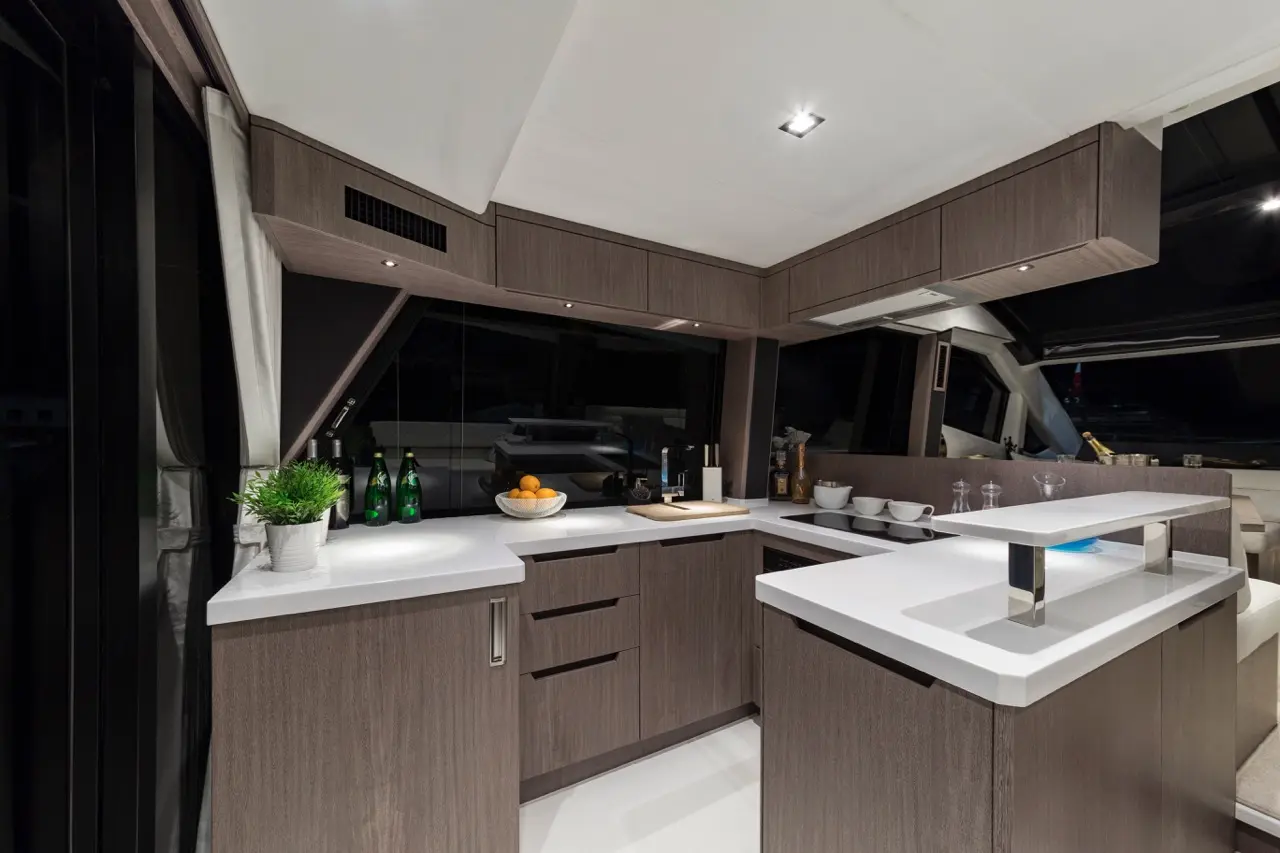 Modern kitchen with dark wood cabinets, white countertops, potted plant, fruit bowl, and glasses, set against large windows.