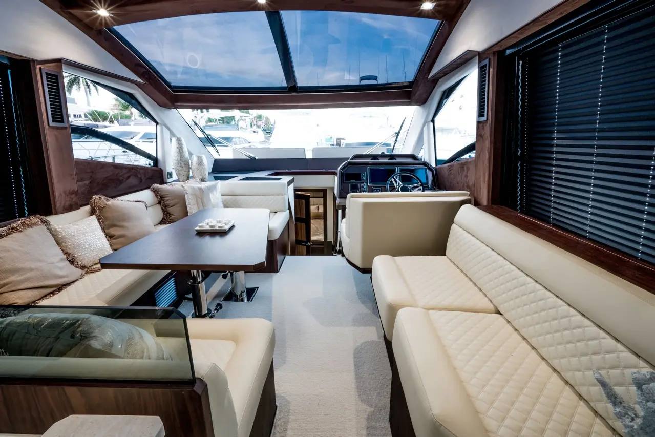 Luxurious yacht interior featuring plush seating, a wooden table, and a panoramic window view of docked boats outside.