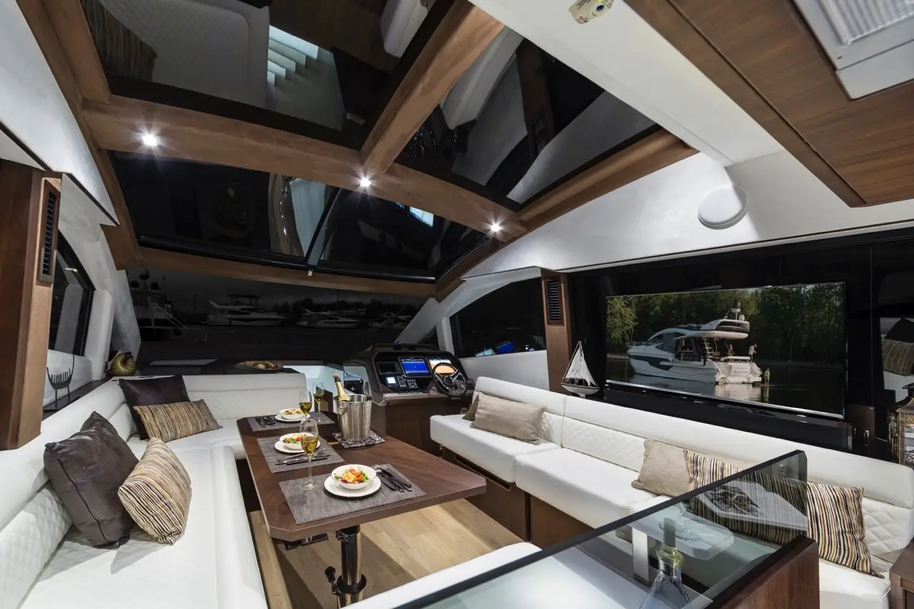 Luxurious boat interior featuring a dining area with food, drinks, sleek furniture, and panoramic windows showing yachts outside.