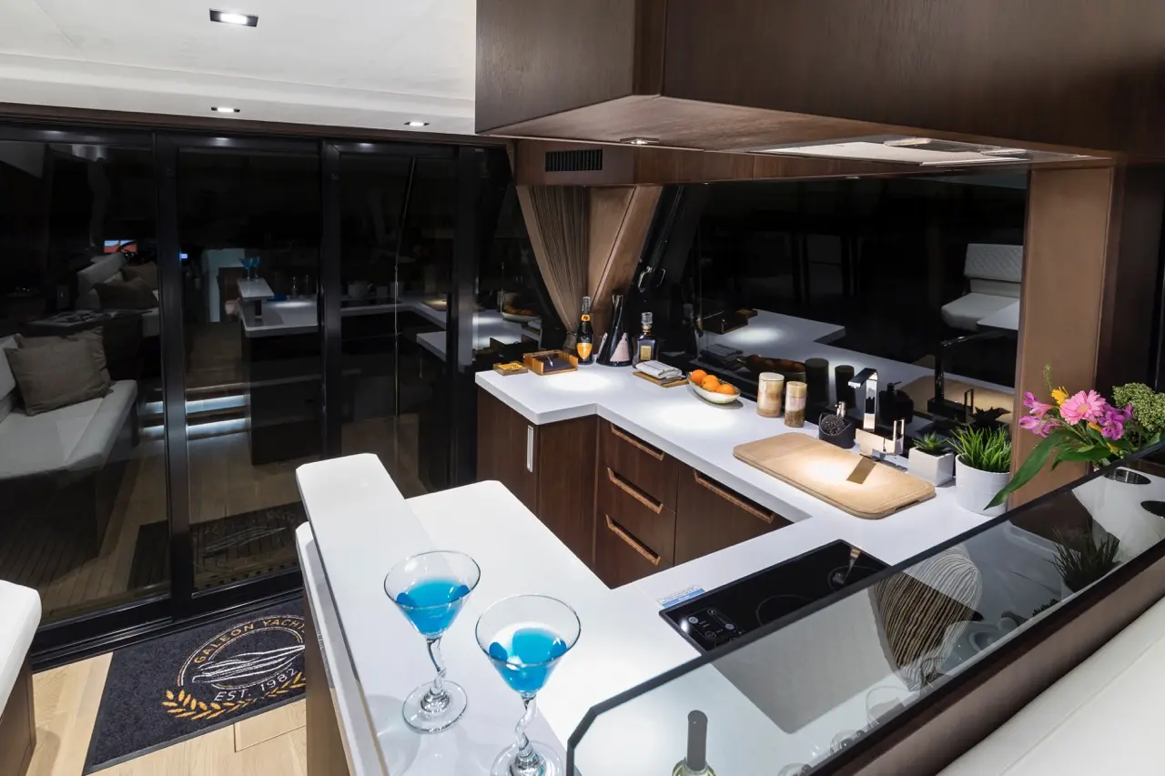 A modern yacht kitchen at night features a sleek design, blue cocktails, fresh fruit, and decorative flowers on the counter.