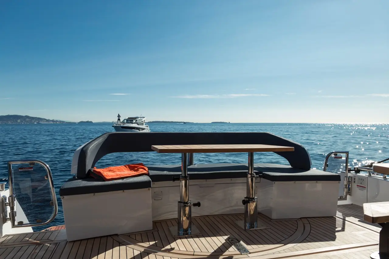 A sleek boat interior features a table and cushioned seating, with a scenic ocean view and another boat in the distance.