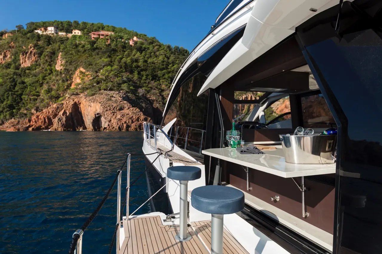 Luxury yacht with a sleek design, featuring stools and a bar area, overlooking a rocky coastline and blue waters.
