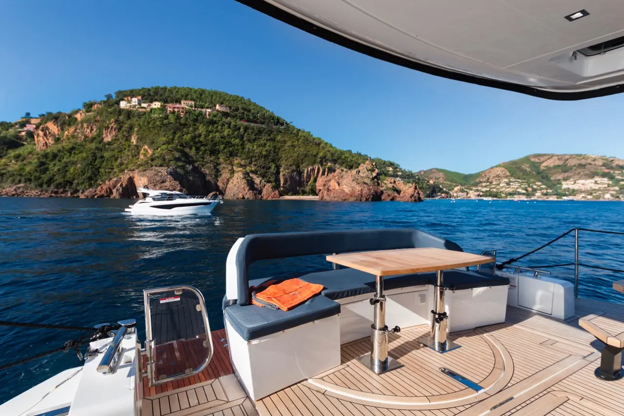 A luxury yacht deck features a wooden table, upholstered seating, and orange towels, with a scenic coastal backdrop.