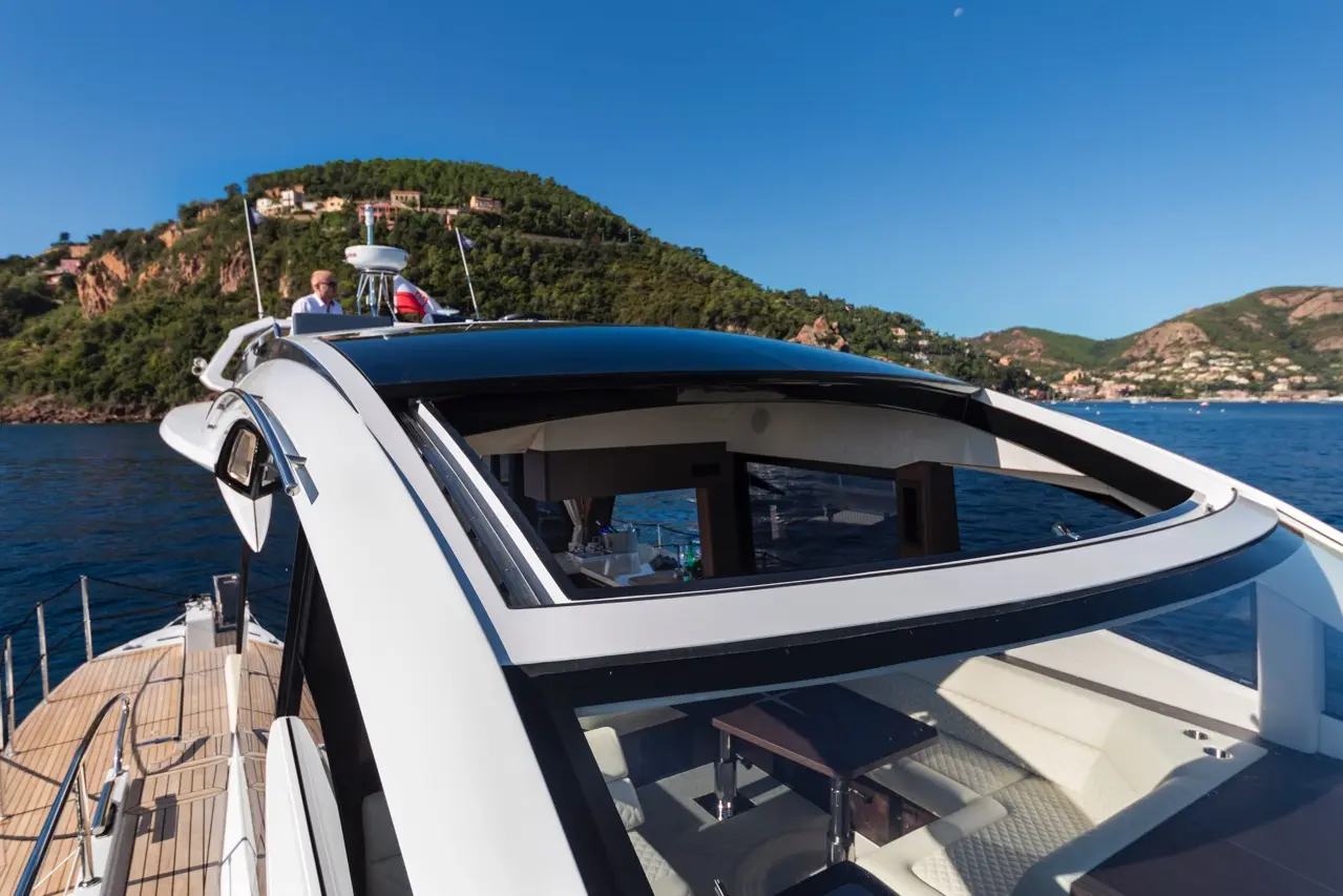 Luxury yacht view with a sunroof, blue water, and lush green hills in the background, featuring a man steering the boat.
