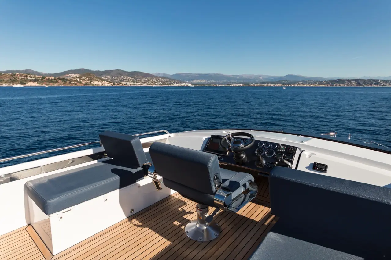 View from a yacht's helm showing the steering wheel, controls, and open water, with a coastline in the background.