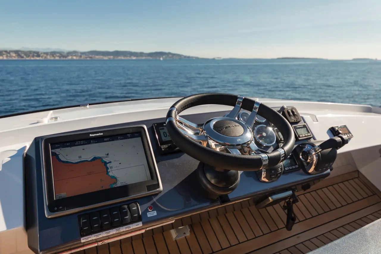 Cockpit view of a yacht featuring a steering wheel, navigation screen showing a map, and a sunny seascape in the background.