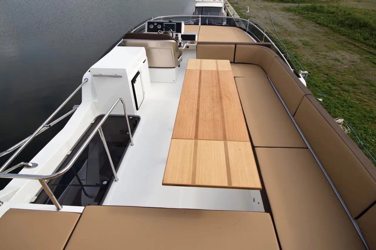 Deck of a boat featuring a wooden table, cushioned seating, and a view of water and grassy area in the background.