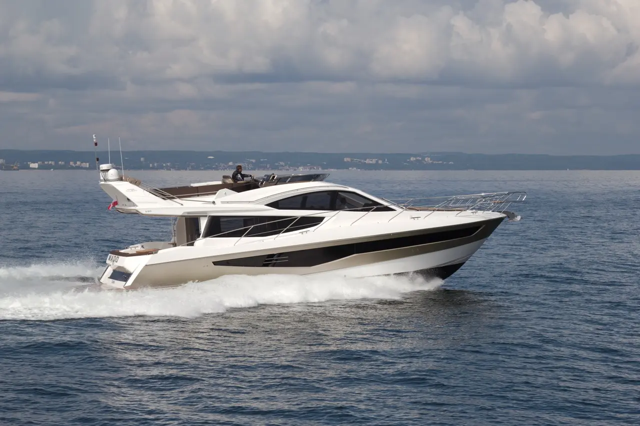 A sleek motor yacht cruises through calm waters, creating white frothy waves, under a partly cloudy sky.