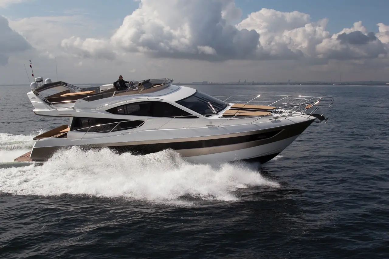 A sleek white yacht is cruising through blue waters, leaving a trail of frothy waves, with a person at the helm.