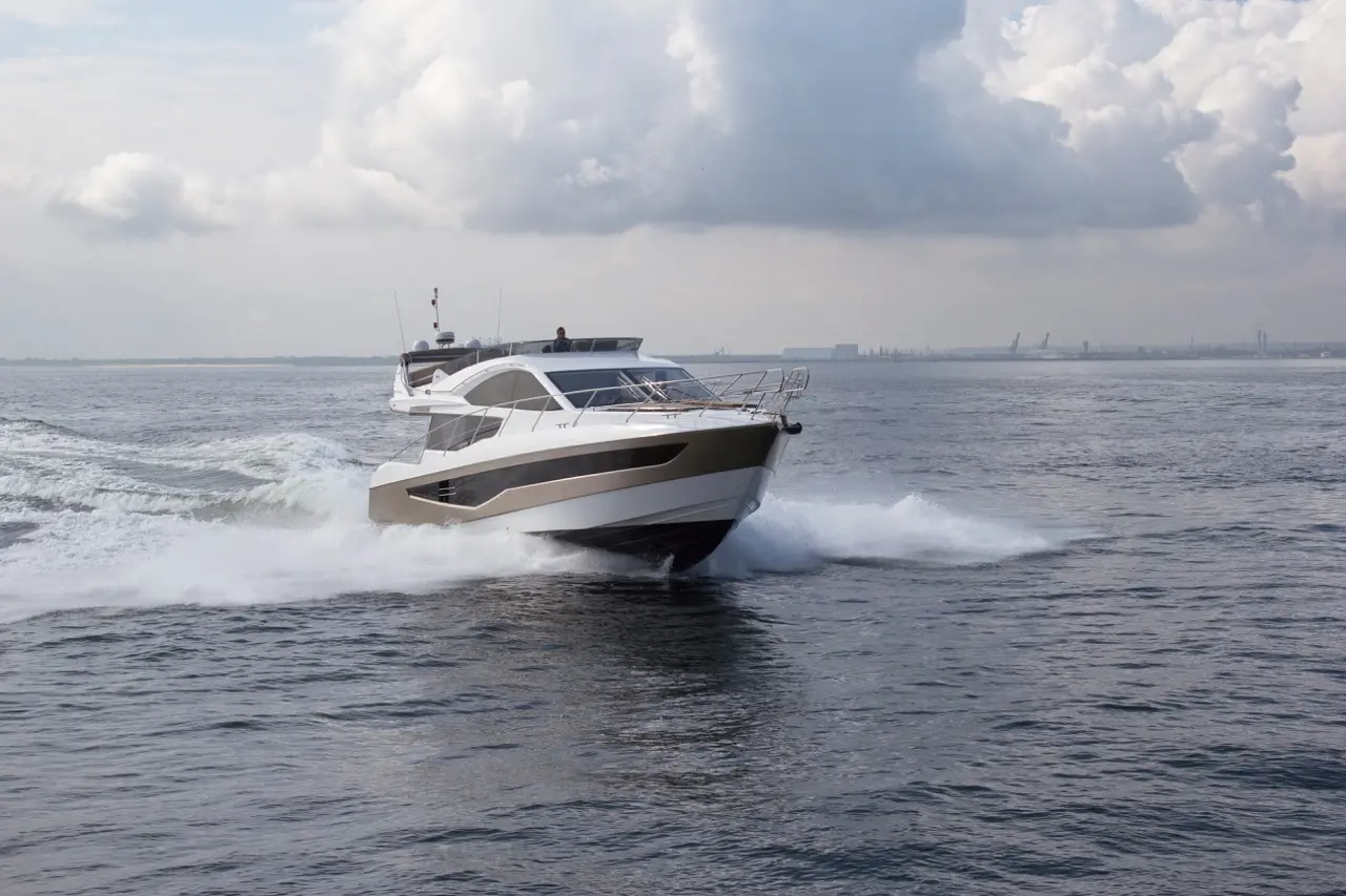 A sleek motor yacht glides over waves, with spray flying, under a cloudy sky on a calm body of water.