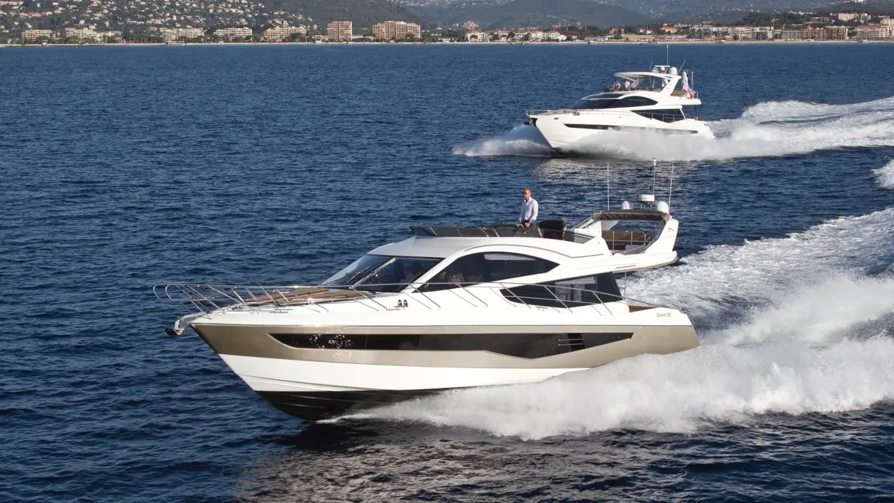 Two luxury yachts speed across a blue ocean, with a coastal city and mountains in the background, showcasing a sunny day.