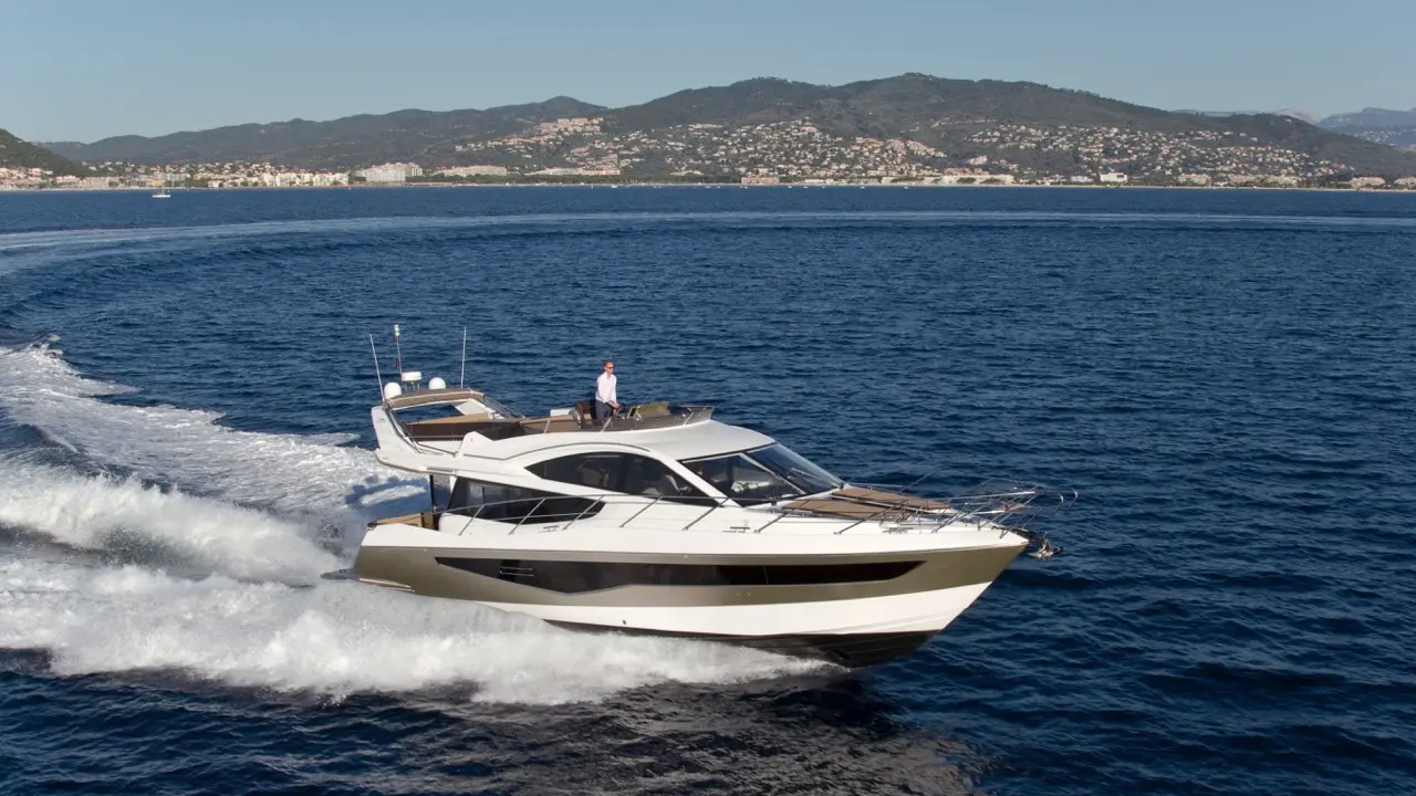 Luxury motor yacht cruising on blue water with a man on the deck, surrounded by hills and shoreline.